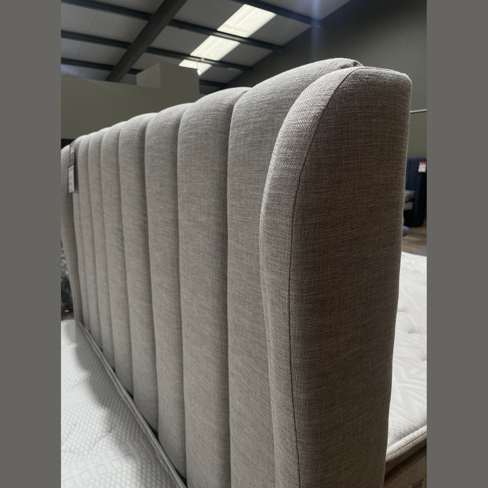A close-up of the Isobella Bedframe’s gray headboard, featuring vertical channel tufting in stone linen fabric, is shown in a showroom with other bedframes and mattresses in the background.