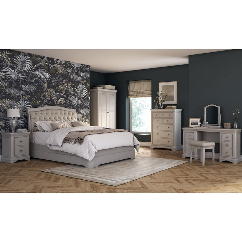 A stylish bedroom with a taupe bed frame, contemporary furniture, and a palm-patterned accent wall. Features a Roman shade, Mabel Bedside tables, dresser, wardrobe, dressing table, wood floor, and rug.