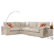 The Dexter Corner is a modern beige corduroy corner sofa, paired with two orange patterned throw pillows, a glass side table with a brown vase, and a curved floor lamp in the background.