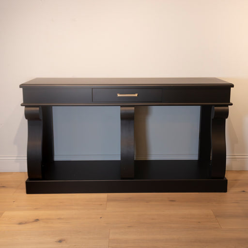 The Large Scroll Console with Drawer features black wood, a single drawer with a silver handle, curved scroll side supports, and an open lower shelf. It stands against a light wall on a wooden floor.