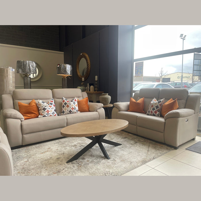 A modern showroom showcases the Lorenzo Electric Recliner Sofa in beige with colorful throw pillows, a wooden oval coffee table, two metallic table lamps, decorative vases, a round mirror, and large windows streaming in natural light.