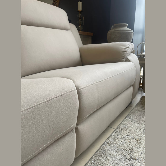 A close-up, angled view of the Lorenzo Electric Recliner Sofa in beige with plush armrests, set on a light tile floor by a textured rug. A large decorative vase and other furniture appear in the background.