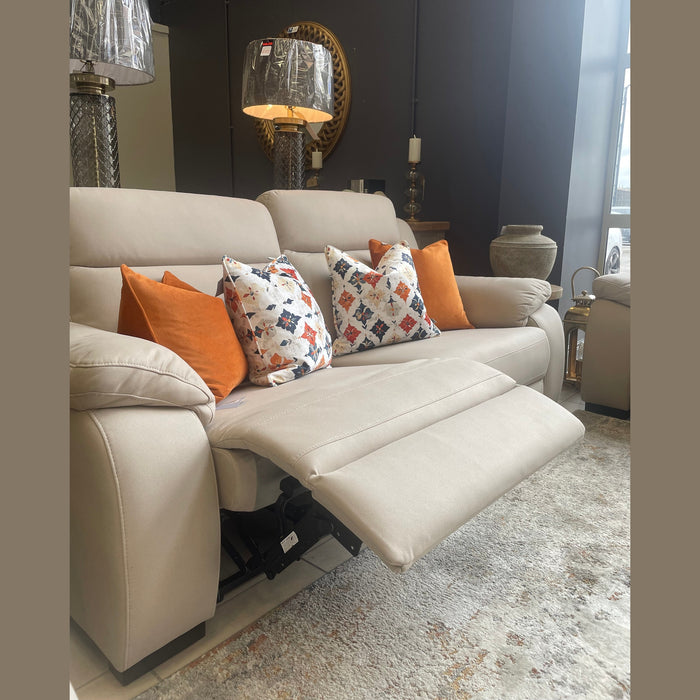 The Lorenzo Electric Recliner Sofa in beige, accented with orange and patterned pillows, is set in a bright living room by the window alongside decorative lamps, a round mirror, and assorted ornaments.