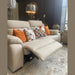 The Lorenzo Electric Recliner Sofa in beige, accented with orange and patterned pillows, is set in a bright living room by the window alongside decorative lamps, a round mirror, and assorted ornaments.