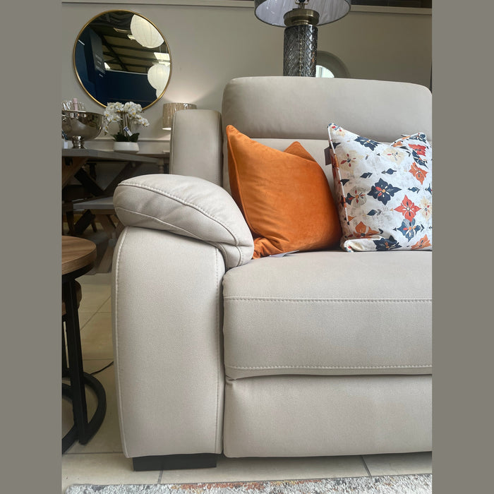 The Lorenzo Electric Recliner Sofa in beige comes with washable fabric, an orange pillow, and a white floral pillow. A round mirror, lamp, and a dining table with flowers are displayed in the background.