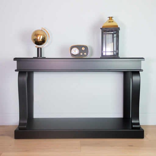 A Scroll console 130cm in black stands against a white wall, displaying a gold globe, a small vintage clock, and a lantern on its bare tabletop. Light wooden flooring completes the look.