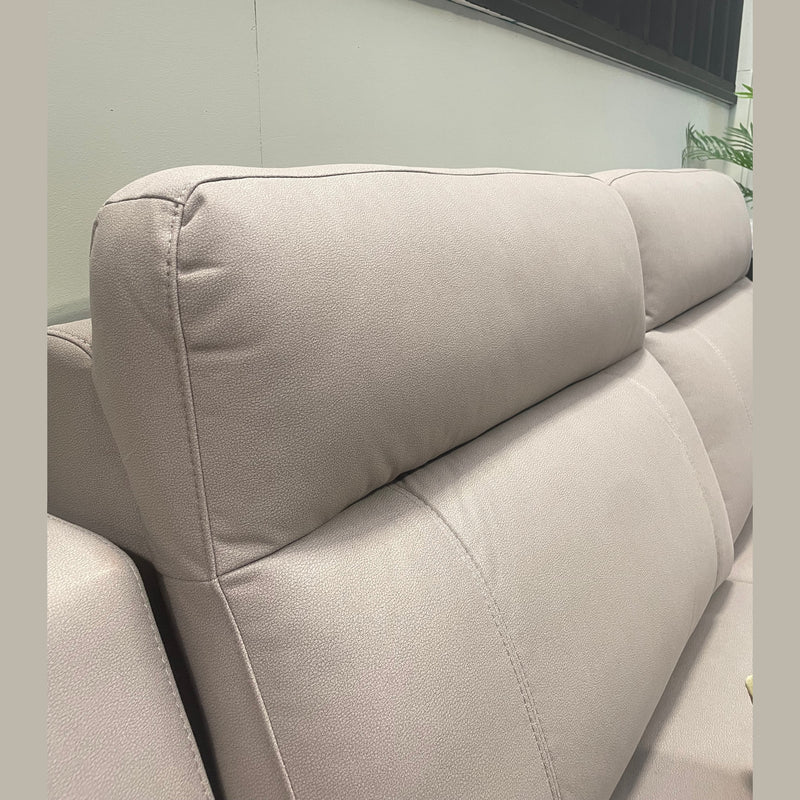 Close-up of the Chloe Electric Recliner, a contemporary taupe microfibre sofa featuring plush, rectangular backrest cushions with visible stitching, set against a pale wall. A touch of green from a plant appears in the background on the right.