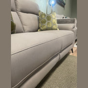 Close-up of the Chloe Electric Recliner in light gray, adorned with a green hexagon-patterned pillow, situated next to a gray couch in a cozy living room with a rug and a glowing lamp in the background.