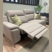 The Chloe Electric Recliner in taupe microfibre, paired with two green geometric-patterned pillows, a lamp, and potted plants, creates a cozy focal point in this modern living room setting.