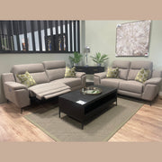 Modern living room with two beige sofas, including the sleek Chloe Electric Recliner featuring a reclined seat. There are green-patterned pillows, a black coffee table, a rug, plants, wall art, and a console table with a lamp and decor.