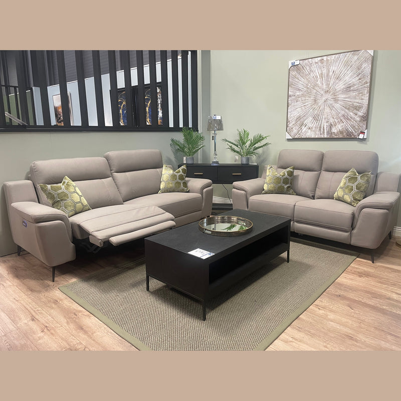 Modern living room with two beige sofas, including the sleek Chloe Electric Recliner featuring a reclined seat. There are green-patterned pillows, a black coffee table, a rug, plants, wall art, and a console table with a lamp and decor.