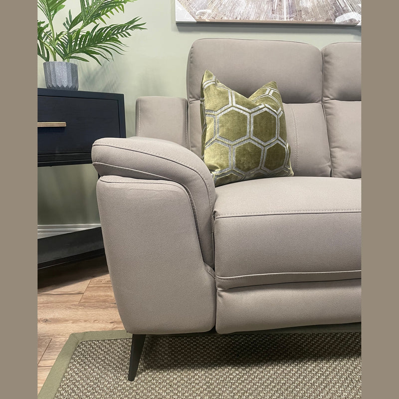 The Chloe Electric Recliner in contemporary taupe microfibre, accented with a green geometric-patterned pillow, sits atop a textured rug. A potted plant and a black side table in the background complete the cozy, modern living room setting.