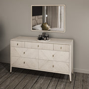 The Diletta Wide 7 Drawer Chest in ebony and sand wood with gold handles stands against the wall, topped with perfume bottles. A matching framed mirror above reflects the bed and curtains, complementing the wood plank flooring.