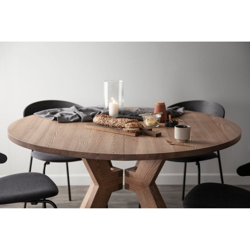 The Monogram Custom Made Table is shown round with four dark chairs, topped with a loaf of bread on a cutting board, a lit glass candle, a mug, small dishes, and draped in a gray cloth against a plain light background.