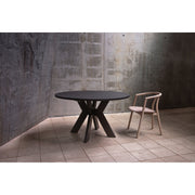 The Monogram Custom Made Table, a modern round ash dining table with an angular base, stands on a tiled floor beside a light wood chair with a curved backrest, set against concrete and textured grey walls.