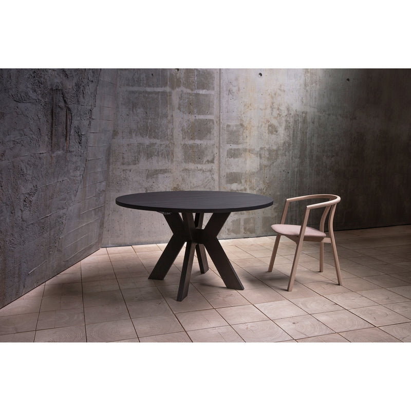 The Monogram Custom Made Table, a modern round ash dining table with an angular base, stands on a tiled floor beside a light wood chair with a curved backrest, set against concrete and textured grey walls.