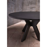 The Monogram Custom Made Table, a modern round dining table with angled legs, stands on light wooden flooring against a textured gray concrete wall.