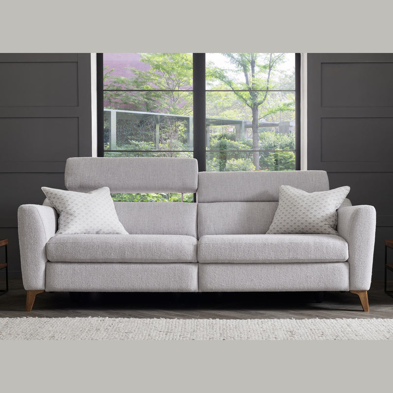 The Otis Sofa Range, upholstered in Aquaclean fabric and paired with two patterned pillows, is placed before a large window overlooking greenery. The setting includes dark paneled walls and a light-colored rug.