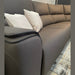 A close-up of the Vegas Corner Group sofa in modern gray, featuring two light beige throw pillows on a patterned rug against a neutral wall with a round mirror above. Its wipeable fabric ensures easy maintenance.