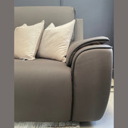 A close-up of the Vegas Corner Group, a modern gray sofa with wipeable fabric and two light beige throw pillows, set against a gray wall. Its textured finish and plush armrest are shown, with part of a patterned rug on the floor.