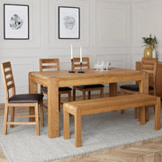 Natural solid oak wood dining set with a rectangular extending table, four ladder-back chairs with dark faux leather seats, and a matching wooden bench.