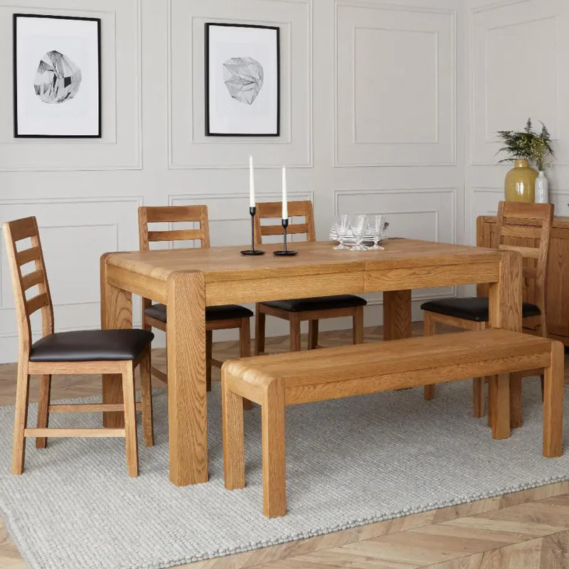 Natural solid oak wood dining set with a rectangular extending table, four ladder-back chairs with dark faux leather seats, and a matching wooden bench.
