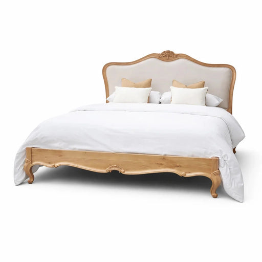 The Toulouse Bedframe features a wooden frame and upholstered headboard, styled with white bedding and four decorative pillows, set against a plain background to evoke classic French oak bedroom elegance.