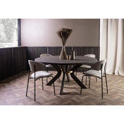 A modern round dining table made of ash wood, with a central decorative vase on the table, surrounded by four chairs with upholstered seats. The table is placed in a room with parquet flooring and a large window.