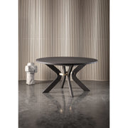 The Sydney Round Dining Table with angled legs stands on a polished concrete floor before vertical beige slats, with a small abstract stone sculpture placed to its left.