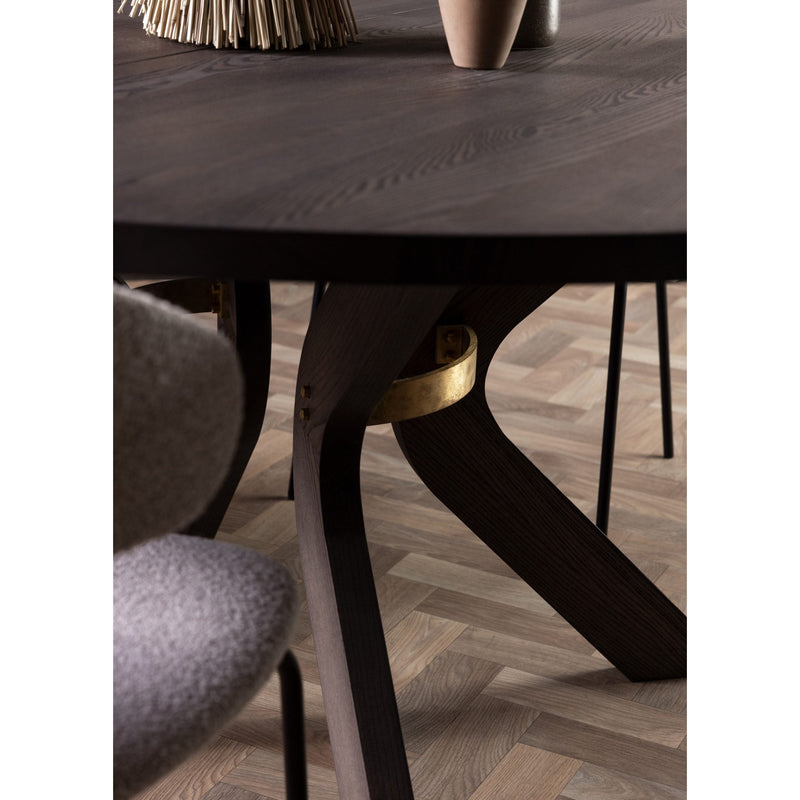 Close-up of the Sydney Round Dining Table in solid ash wood, featuring extendable design and angled legs with a brass accent, set on a light herringbone wood floor. A textured chair and decorative accents are partially visible.