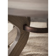 Close-up of the Sydney Round Dining Table leg, crafted from solid ash with a dark bronze metal support bar, resting on a textured beige rug. The natural wood grain highlights its elegance and durability.
