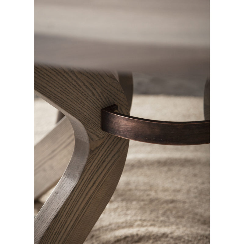 Close-up of the Sydney Round Dining Table leg, crafted from solid ash with a dark bronze metal support bar, resting on a textured beige rug. The natural wood grain highlights its elegance and durability.