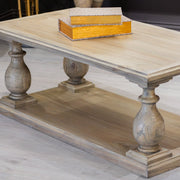 The Marwood Coffee Table features light wood, two ornate carved pedestal legs, and a lower shelf. A golden rectangular tray with books rests on top. Measuring 120 cm wide and 60 cm deep, it's perfect for spacious living rooms.