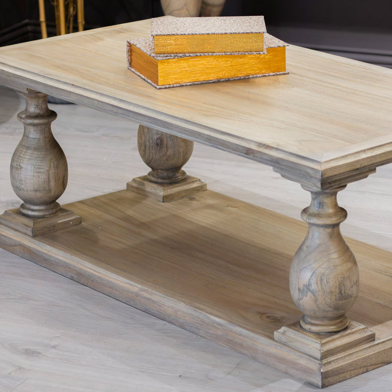 The Marwood Coffee Table features light wood, two ornate carved pedestal legs, and a lower shelf. A golden rectangular tray with books rests on top. Measuring 120 cm wide and 60 cm deep, it's perfect for spacious living rooms.