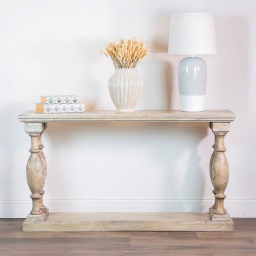 The Marwood Large Console Table (82 cm high, 150 cm wide) features turned legs and displays a white vase of wheat, stacked books, and a white ceramic lamp against a light wall and wooden floor.