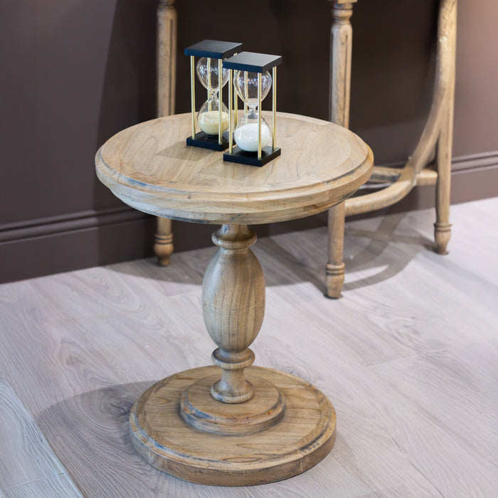 The Marwood Round Side Table (50 cm wide, 57 cm high) with a pedestal base stands on a light wood floor, displaying two black-framed hourglasses. A wooden chair is partially visible in the background.