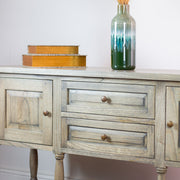 The Marwood Large Sideboard Console, measuring 82 cm high and 150 cm wide with two drawers and cabinet doors, is topped with a tall glass vase and two stacked orange-gold boxes, set against a white wall.