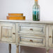 The Marwood Large Sideboard Console, measuring 82 cm high and 150 cm wide with two drawers and cabinet doors, is topped with a tall glass vase and two stacked orange-gold boxes, set against a white wall.