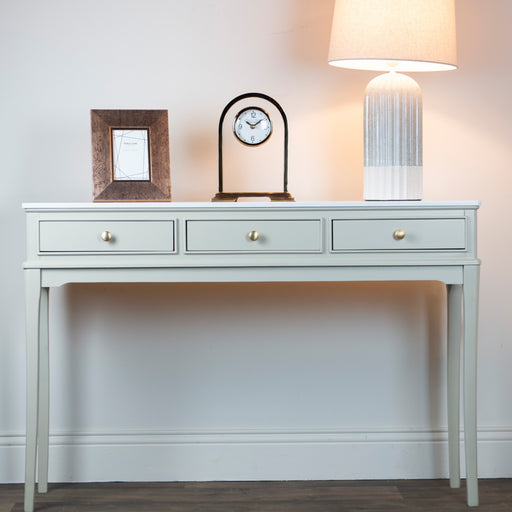 The Monroe Large Console - Stone, featuring three drawers and a marble top, displays a picture frame, round clock, and textured white table lamp against a light wall and wood floor.