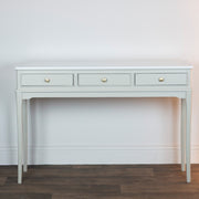 The Monroe Large Console - Stone features a light gray finish, three drawers with round gold knobs, and slender legs—an elegant storage piece that complements any space against a white wall and dark wood floor.