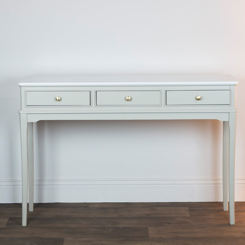 The Monroe Large Console - Stone features a light gray finish, three drawers with round gold knobs, and slender legs—an elegant storage piece that complements any space against a white wall and dark wood floor.