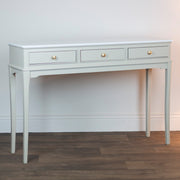 The Monroe Large Console - Stone features a light gray finish, three drawers with round gold knobs, and four slender legs. Elegant and modern, it stands against a white wall on a wood floor.