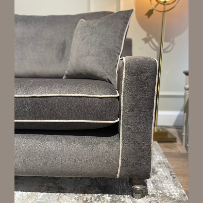 A close-up of the modern grey Emperor Range suite with white piping, featuring one armrest and a cushion. In the background, a gold floor lamp with a round shade and part of a small side table sit on a patterned rug.