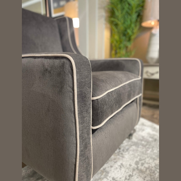 Close-up of an Emperor Range gray upholstered armchair, made from Aqua Clean fabrics with light piping, set on a patterned rug. A potted plant and lamp appear blurred in the background.