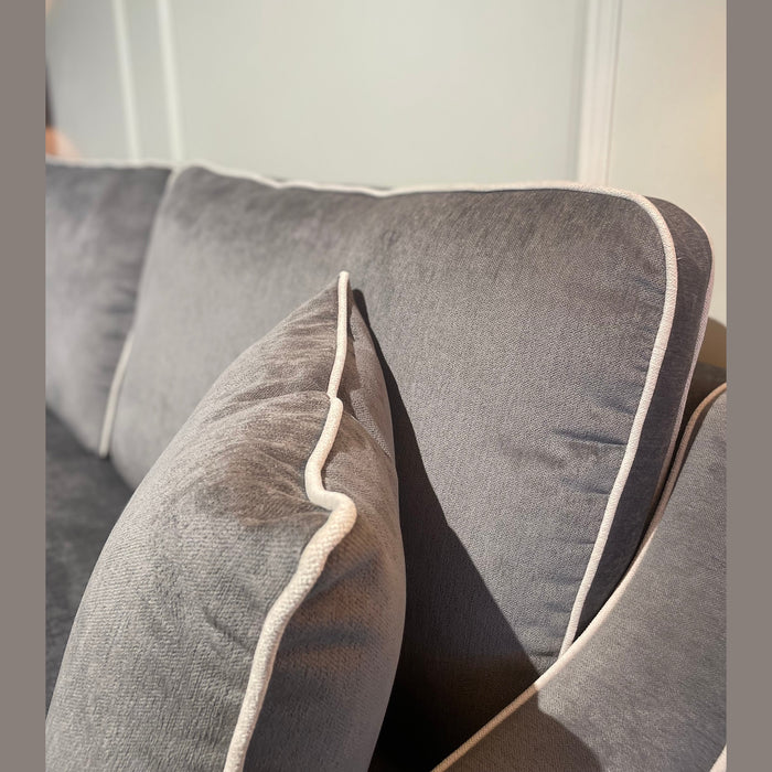Close-up of a grey Emperor Range sofa with white piping and a matching cushion. Sunlight highlights the Irish-made suite fabric's texture, with part of a framed artwork visible on the wall in the background.