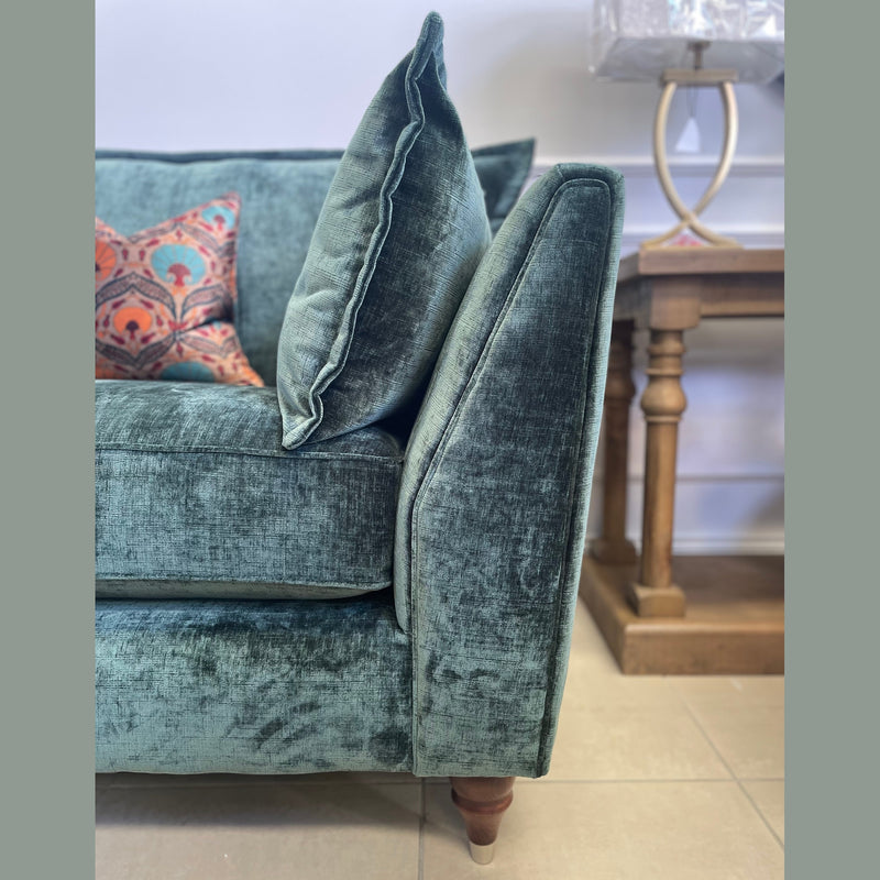 A close-up of the Cleo Range green velvet sofa with deep seats and wooden legs, shown with a matching cushion, a colorful pillow, a wooden side table, and a decorative lamp in the background.