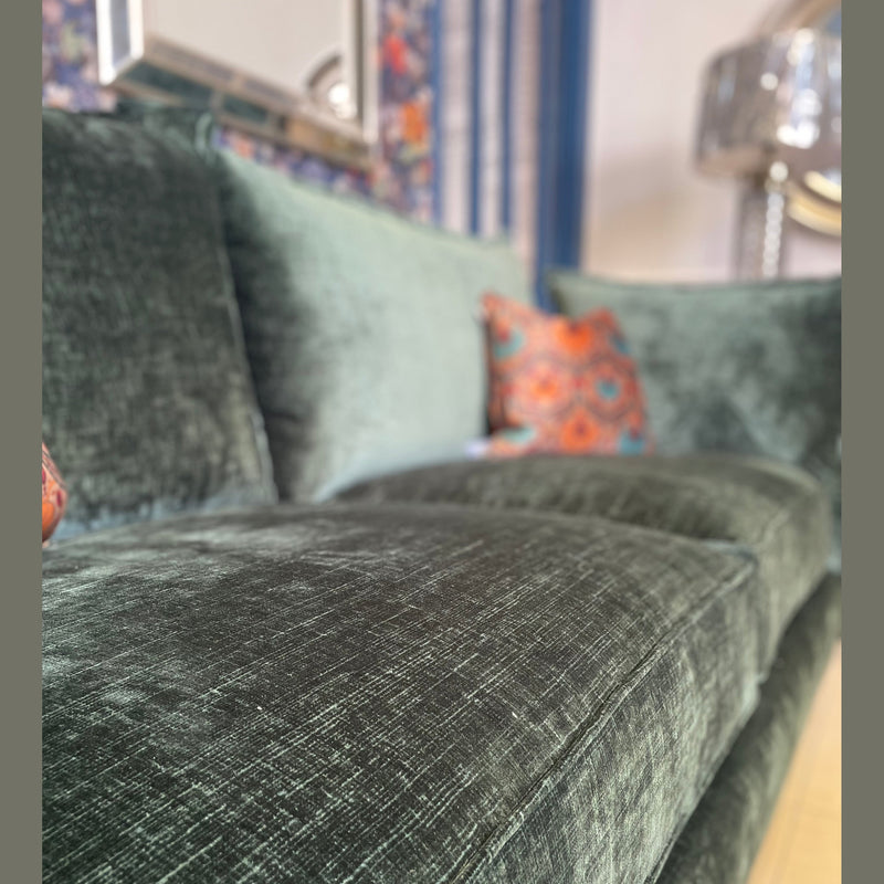 Close-up of the Cleo Range: a plush, textured green velvet sofa with deep seats and a colorful patterned cushion. Elegant style is highlighted, with a softly blurred patterned wall, mirror, and metallic lamp in the background.