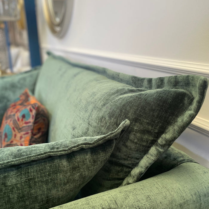 A close-up of a green Cleo Range sofa with deep seats and plush cushions, accented by a colorful patterned pillow. A round mirror and part of a shelf appear blurred in the background.