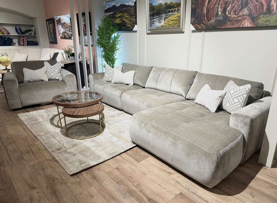 A modern living room featuring the Opus Chaise in a light gray modular design, matching armchair, Aquaclean Fabrics pillows, round wooden coffee table, beige rug, indoor plant, and landscape paintings on the wall.
