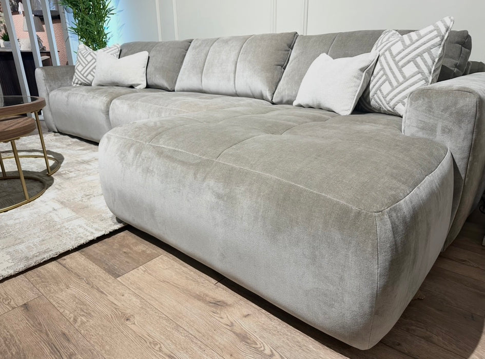 The Opus Chaise is a spacious, light gray sectional sofa with plush cushions in Aquaclean Fabrics, featuring a contemporary design and accented with patterned and solid throw pillows. It’s paired with a round coffee table on a beige rug.